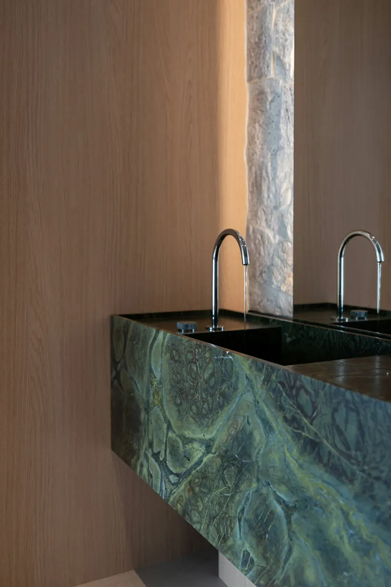 Two arched chrome faucets over a green marble basin, set against warm oak paneling